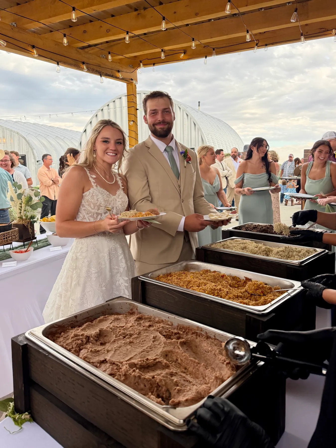 A bride and groom filling their plates at the buffet as guests line up behind them.