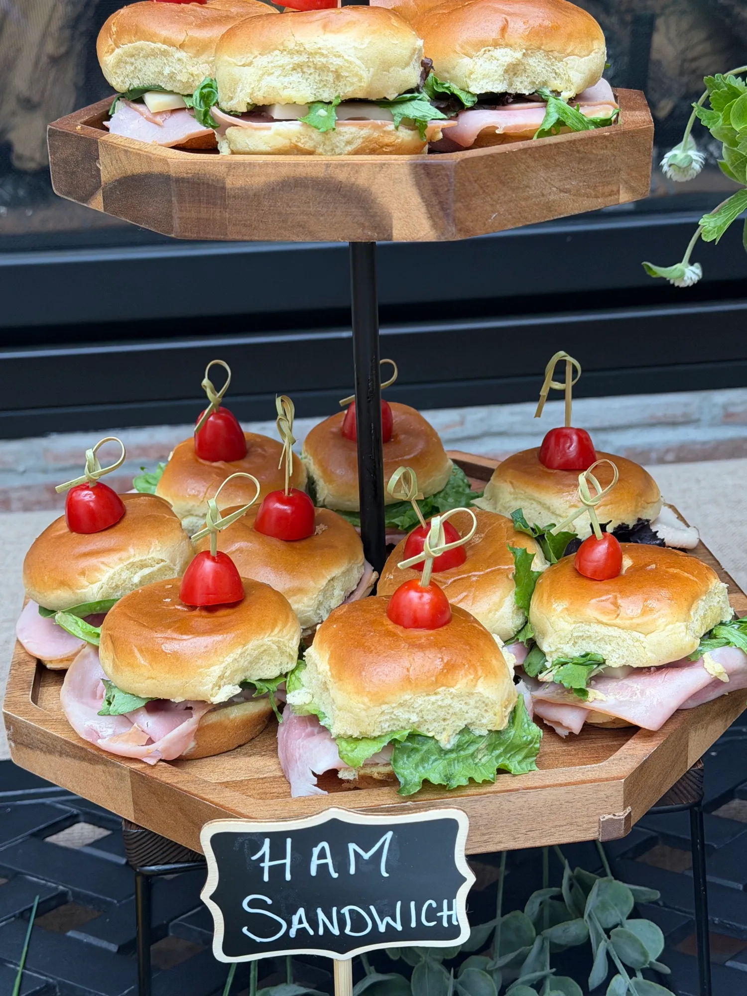 Ham sliders on a tiered wooden stand, each pinned with a cherry tomato.