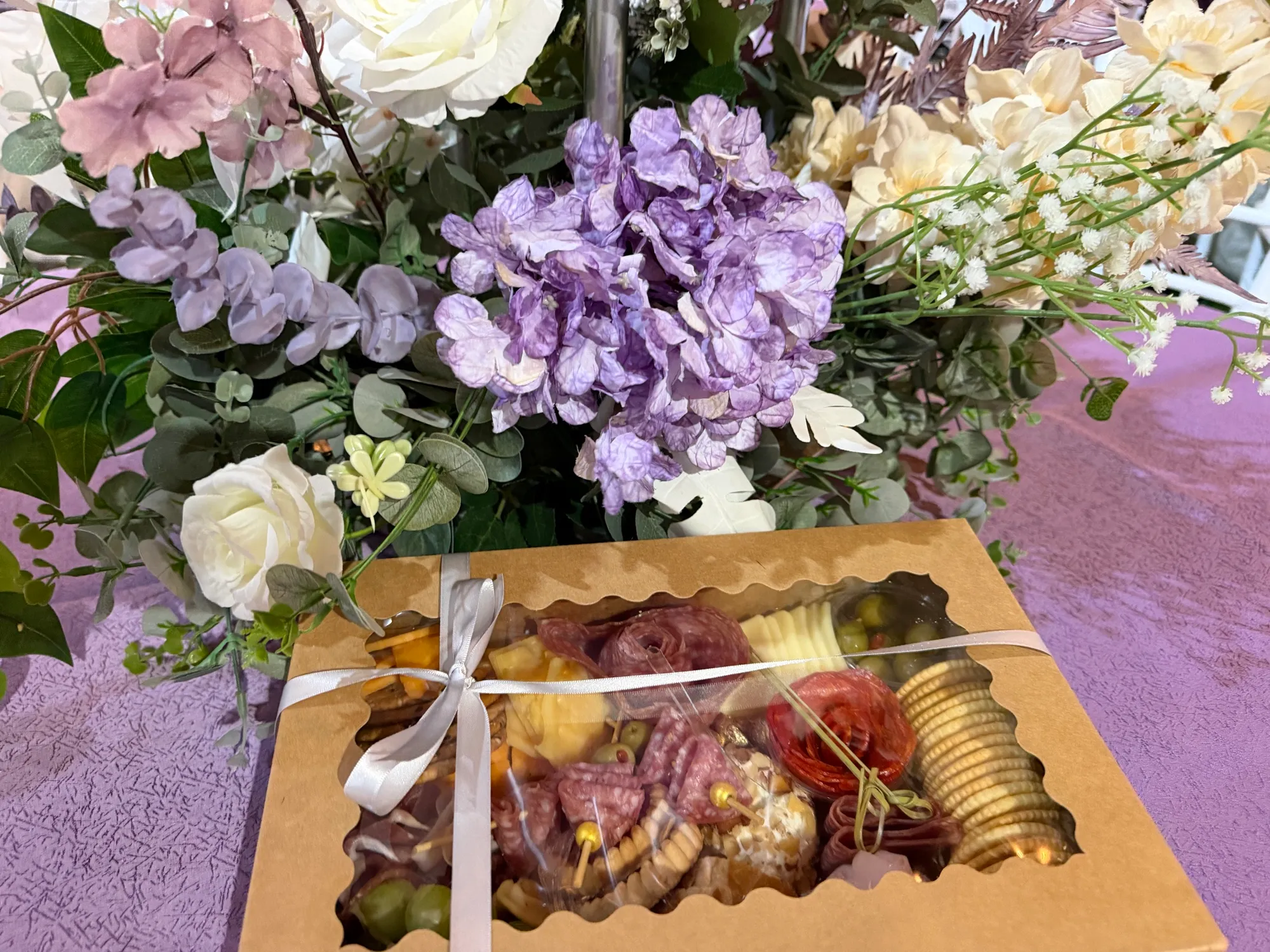 An individual charcuterie box tied with ribbon, set beside fresh flowers.